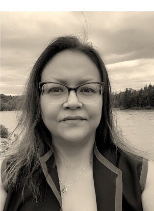 Portrait of Lynn Wright with long dark hair and glasses, wearing a dark vest, standing by a river with trees and a cloudy sky behind her